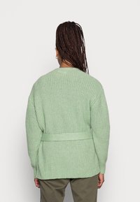 Jasnozielony, teksturowany kardigan z wiązanym w pasie fasonem i luźnym krojem. Posiada szerokie rękawy oraz wzór z ribbingu. Zaprezentowany widok z tyłu.