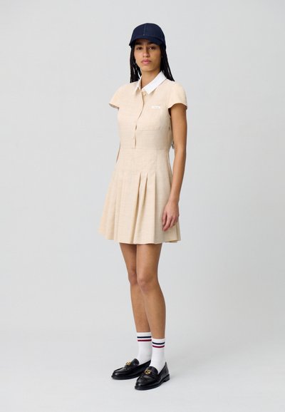 Robe beige à manches courtes avec un devant à boutons, un col et deux poches latérales ; design de jupe plissée. Portée avec une casquette bleu marine et des mocassins noirs.