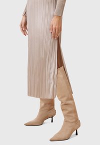 Abito midi plissettato in morbido tessuto beige, con uno spacco laterale. Abbinato a stivali al ginocchio in suede beige chiaro con punta affilata e tacco a stiletto.