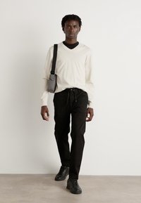 Witte trui met lange mouwen, zwarte joggingbroek met trekkoord en zwarte sneakers. Een grijze crossbody tas is over de schouder gedragen. Effen achtergrond.