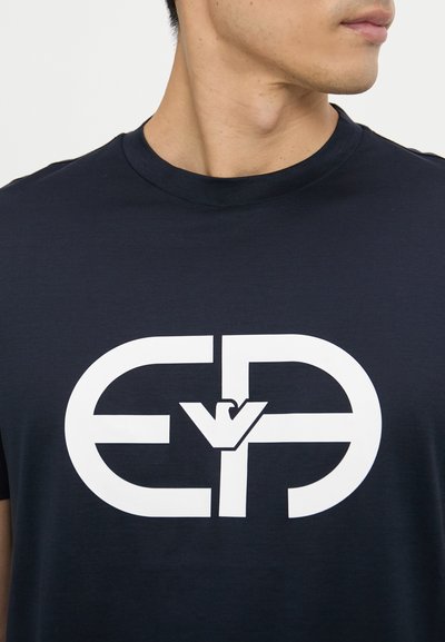 Tricou din bumbac albastru închis, având un mare logo grafic alb în centru, cu guler rotund și mâneci scurte.