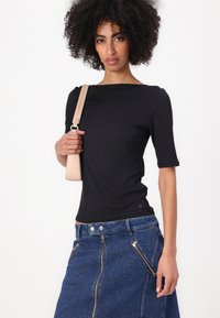 Svart ribbet topp med ermelengde til albuen, kombinert med en blå denimskjørt med doble glidelåser og høy midje. Holder en beige skulderveske.