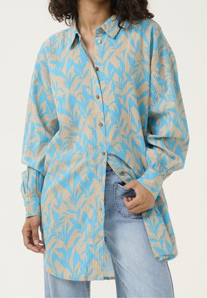 Femme portant une longue chemise beige et bleue à motifs de feuilles, boutonnée, sur un jean bleu clair, avec une main dans une poche.