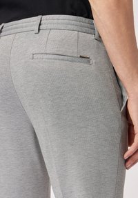 Grijze broek van gestructureerde, gebreide stof met een ritssluiting achterzak en subtiele taillebanddetails, weergegeven vanuit een zijaanzicht.