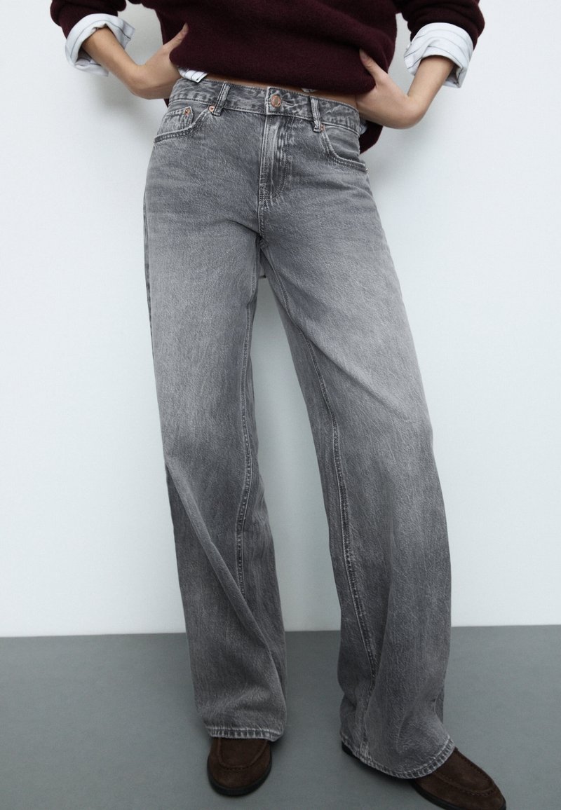 Personne portant un jean gris délavé et ample, des chaussures marron, les mains sur les hanches, avec un pull bordeaux et des manches de chemise blanche retroussées.