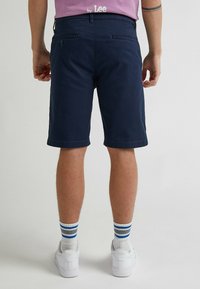 Pantaloni corti blu navy in misto cotone, con tasche posteriori e orli arrotolati; indossati con scarpe da ginnastica bianche e calzini a righe.
