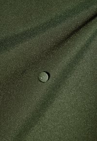Tissu vert foncé affichant une surface texturée, comportant un petit bouton brillant avec un motif subtil, ajoutant du détail au matériau.