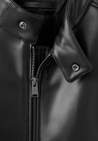 Gros plan du col et de la fermeture éclair d'une veste en cuir noire avec des boutons-pression métalliques visibles et des coutures détaillées.