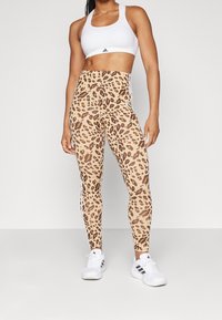 Bruna leopardmönstrade leggings med hög midja, vita sidosträck och en mjuk tygtextur, kombinerat med en vit sport-BH och sneakers.