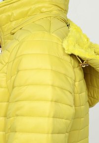 Chaqueta acolchada amarilla con textura acolchada, con ribete de capucha de piel sintética, tela brillante y cremallera inclinada en el hombro.