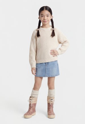 Jauna mergina su pintais plaukais, vilkinti kreminio spalvos megztinį, džinsinę sijoną, storas kojines ir raštuotus žieminius batus, stovinti su ranka ant klubo.