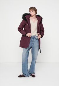 Parka bordeaux avec une capuche en fausse fourrure, fermeture à zip, deux poches latérales, associé à un pull beige et un jean clair évasé.