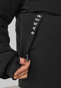 Zwarte pufferjas met een zachte textuur. Voorzien van een opvallende ritstreker met het merk "BAKER" en elastische manchetten voor een nauwsluitende pasvorm.