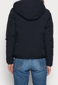 Marineblå pufferjakke med hætte, med kontrastfarvet logobånd på kraven og elastisk kant. Bærers med lyseblå jeans, set bagfra.