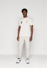 T-shirt cinzenta clara com logótipo cor de vinho e número 10, combinada com calças de treino cinzento claro e ténis preto e branco. Design simples e corte relaxado.