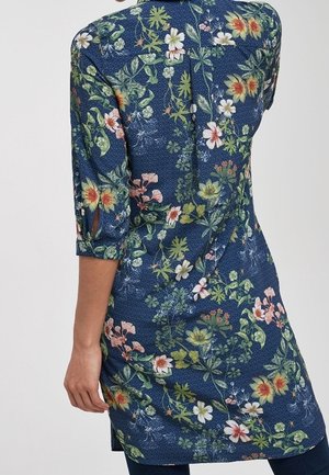 Vue arrière d'une personne portant une robe bleue fleurie longueur genou avec des manches trois-quarts et une texture subtile à pois.