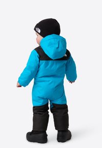 Blå barneute-dress med svarte detaljer, hette og elastiske mansjetter, kombinert med en svart strikkelue. Vanntett materiale og isolert design.