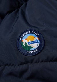 Tummansininen kangas, jossa on brodeerattu pyöreä päällyste. Päällä on vuoria, aurinko ja tekstit "POLARN O PYRET SWEDEN" ja "SINCE 1976."