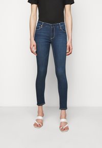 Úzké džíny v tmavě modrém denimu s hladkým povrchem, design s pěti kapsami, přední knoflík a mírně roztrhaný lem. V kombinaci s bílými sandály.