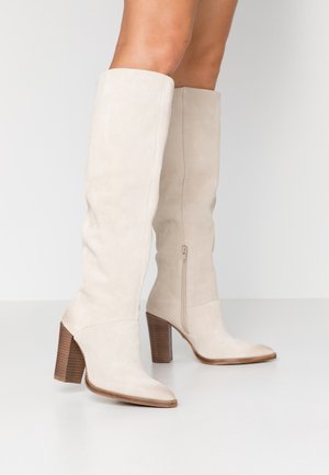 Beige suede knee-high boots with a pointed toe, zip closure, and stacked wooden heel. Smooth texture and sleek design.