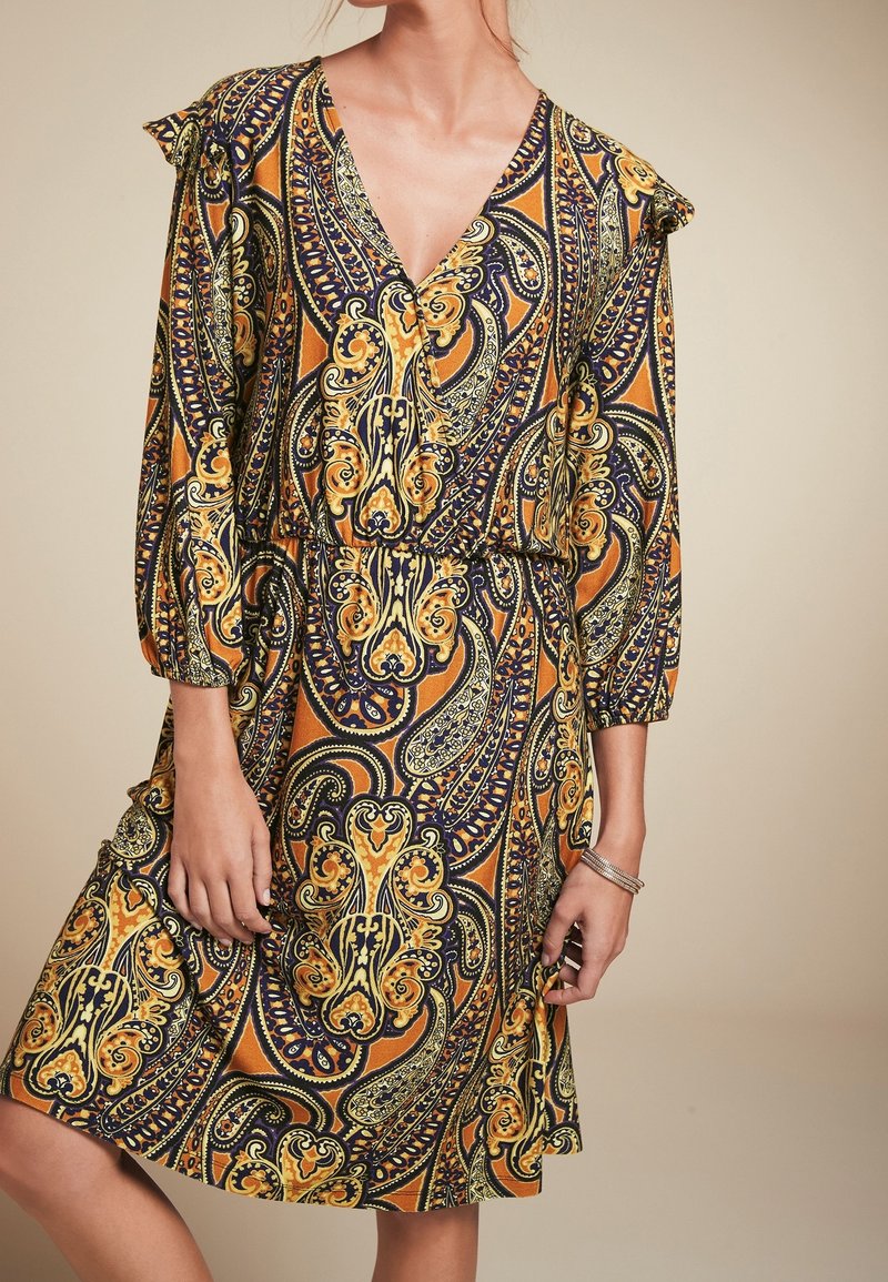Femme portant une robe mi-longue avec des motifs paisley intricats orange, jaune et noir et des manches trois-quarts, se tenant avec une jambe fléchie.