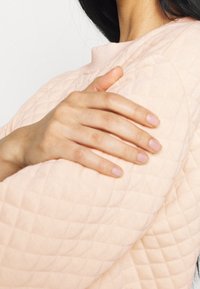 Rosa gesteppter Stoff mit einem strukturierten Muster, der teilweise einen Arm bedeckt. Eine Hand liegt sanft auf dem Stoff und zeigt gepflegte Nägel.