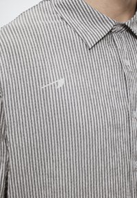 Κοντινό πλάνο ενός ατόμου που φοράει ένα λευκό και γκρι κατακόρυφα ριγέ πουκάμισο με κουμπιά, με αιχμηρό γιακά και μικρό τσεπάκι στο στήθος.