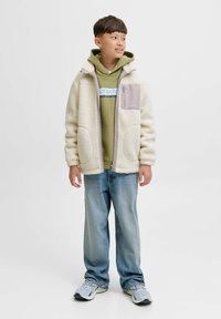Fleece jack in crème met een grijze zak, gedragen over een groene hoodie, gecombineerd met lichtblauwe jeans en grijze sneakers.
