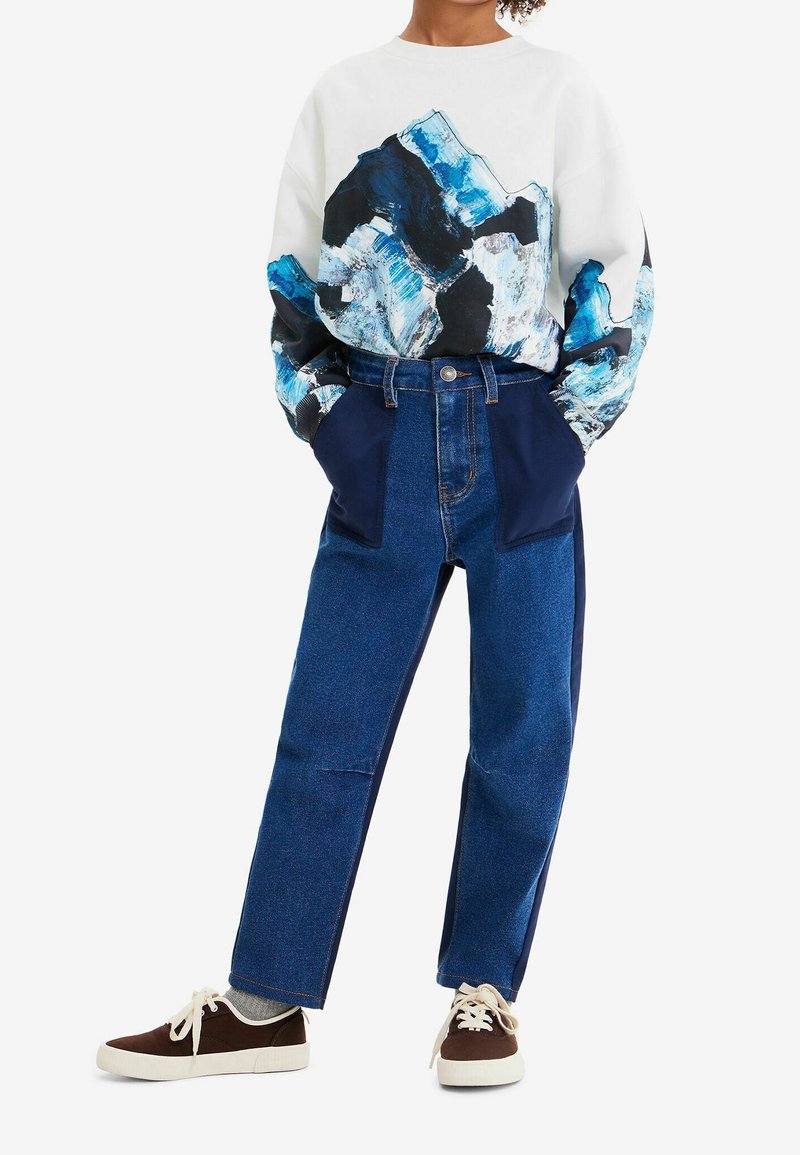 Sudadera blanca con un diseño abstracto de montaña en azul y negro. Combinada con pantalones de mezclilla azul con acentos en azul oscuro y zapatillas marrones.