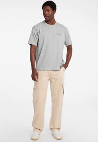 Sivé tričko s krátkym rukávom a jemným logom na hrudi, v kombinácii s béžovými cargo nohavicami s viacerými vreckami a voľným strihom.