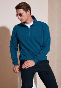 Türkisfarbener Quarter-Zip-Pullover aus weichem Stoff mit langen Ärmeln, ausgestattet mit einem horizontalen Nahtdetail und über einem weißen Shirt getragen.
