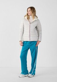 Chaqueta acolchada gris claro con capucha, con un zipper rojo y azul. Pantalones de chándal azul verdoso con rayas laterales blancas, complementados por zapatillas plateadas.