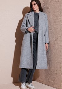 Lichtgrijze wollen blendjas met een lange, op maat gemaakte silhoutte, brede revers en dubbele knoopsluiting, gecombineerd met een grijze trui en donkere jeans.