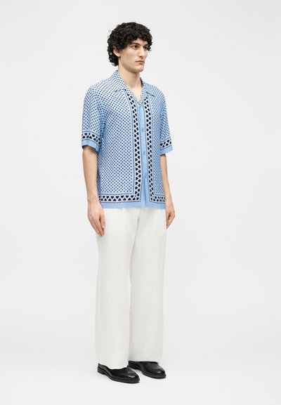 Άνδρας με σγουρά μαλλιά, φοράει ένα μπλε πουκάμισο με σχέδια και κοντά μανίκια, λευκό παντελόνι φαρδύ και μαύρα παπούτσια, στέκεται μπροστά σε απλό φόντο.