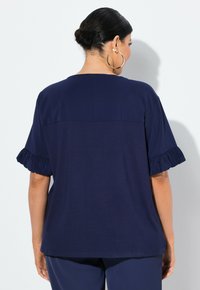 Top blu navy a maniche corte con maniche a volant, texture liscia e orlo dritto. Presenta un pannello di design a contrasto sul retro.