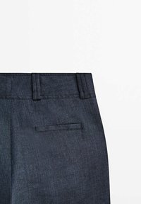 Détail d'un pantalon en denim bleu foncé montrant la taille avec passants de ceinture et une seule poche arrière horizontale sur fond blanc.