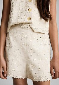 Set di top corto e pantaloni a shorts ricamati beige, caratterizzati da motivi floreali, paillettes, bordi ondulati e bottoni tondi sul top.