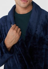 Tmavě modrý plyšový župan s šálovým límcem, měkkou texturou a dlouhými rukávy, nošený přes tmavě zelenou košili. Ruka držící límec.