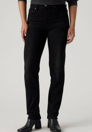 Person iført sorte jeans med lige ben og forlommer samt sorte ankelstøvler med hæl, stående mod en ensfarvet baggrund.