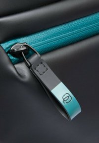 Zwarte ritssluiting op een gestructureerd oppervlak, met een blauwgroene toets en een rechthoekige tag met reliëfdesign, tegen een gladde zwarte achtergrond.