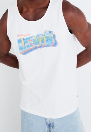 Homme portant un débardeur blanc avec un logo coloré « Greetings from Levi's » et un jean en denim bleu clair, debout devant un fond uni.