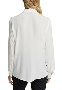 Femme portant une blouse blanche à manches longues avec un col, vue de dos, portée avec un pantalon noir.
