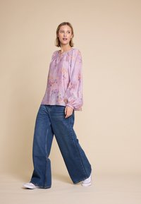 Lys lilla bluse med blomster mønster, elastisk halsudskæring og sammensamlede ærmer, parret med viddebenet blå denim jeans og hvide sneakers.