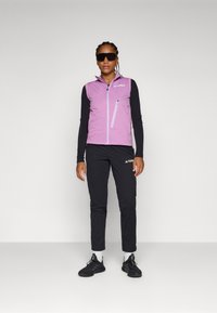 Vesta fără mâneci de culoare violet deschis, cu guler înalt și buzunar cu fermoar, purtată cu pantaloni negri și pantofi sport negri. Ochelari de soare negri purtați.