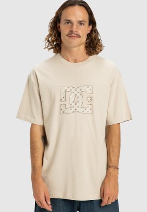 Uomo con i capelli ricci fino alle spalle che indossa una t-shirt oversize beige con un logo DC a fantasia e jeans blu.