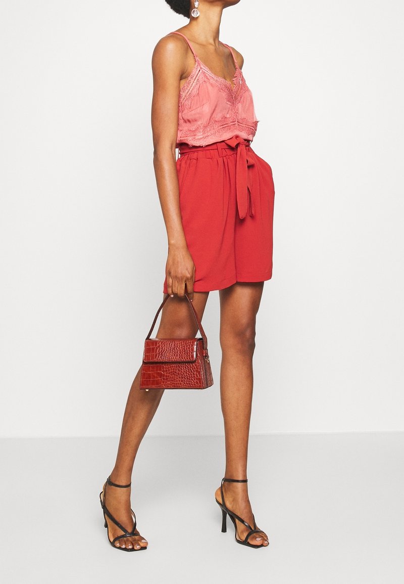 Un short rouge avec une ceinture à nouer, associé à un débardeur en dentelle rose. Des talons aiguilles noirs à lanières et un petit sac à main en croco marron viennent parfaire le look.