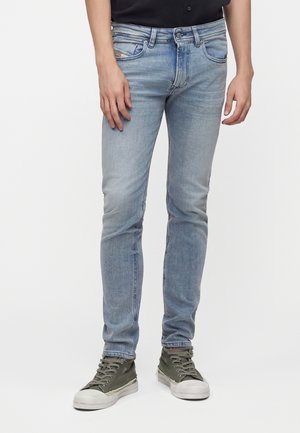 Jean slim bleu clair porté avec des baskets montantes vert olive sur une personne debout sur un fond blanc.