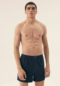 Czarne bawełniane bokserki z elastycznym pasem i dwoma zapięciami na guziki z przodu, modelowane przez osobę bez koszuli.