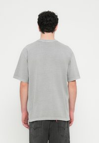 Man with curly dark hair wearing gray short-sleeve t-shirt and black jeans, facing away against a white background.