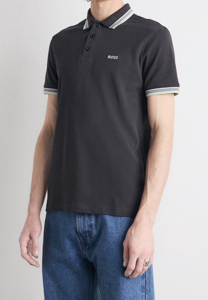 Polo gris foncé avec un col et des poignets à rayures contrastantes vertes et blanches, présentant un petit logo brodé sur la poitrine.